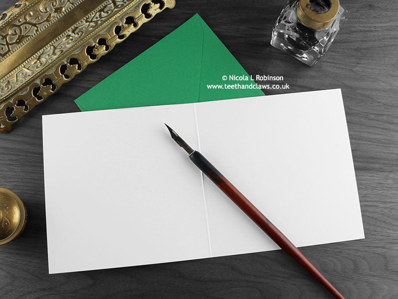 Dinosaur birthday card, shown open and flat with blank inside, on a green envelope, alongside vintage Victorian pen and writing desk accessories. Design and illustration © Nicola L Robinson | Teeth and Claws