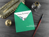 Dinosaur birthday card, shown lying flat, with my original pen and ink illustration of a Pteranodon, inside an open green envelope. With vintage Victorian pen and writing desk accessories. Design and illustration © Nicola L Robinson | Teeth and Claws