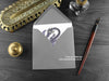 Purple Flying Dragon Greeting Card, shown lying flat, with my original pen and ink illustration of a purple dragon, inside an open grey envelope. With vintage Victorian pen and writing desk accessories. Design and illustration © Nicola L Robinson | Teeth and Claws