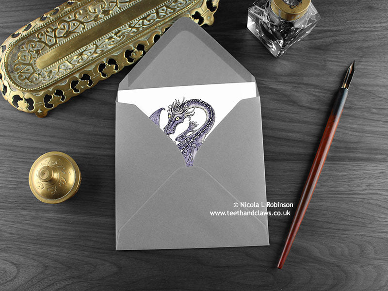 Purple Flying Dragon Greeting Card, shown lying flat, with my original pen and ink illustration of a purple dragon, inside an open grey envelope. With vintage Victorian pen and writing desk accessories. Design and illustration © Nicola L Robinson | Teeth and Claws