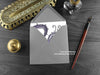 Gothic Purple Dragon Greeting Card, shown lying flat, with my original pen and ink illustration of a purple dragon, inside an open grey envelope. With vintage Victorian pen and writing desk accessories. Design and illustration © Nicola L Robinson | Teeth and Claws