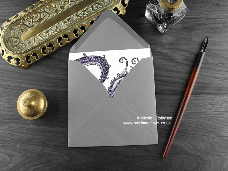 Gothic Purple Dragon Greeting Card, shown lying flat, with my original pen and ink illustration of a purple dragon, inside an open grey envelope. With vintage Victorian pen and writing desk accessories. Design and illustration © Nicola L Robinson | Teeth and Claws