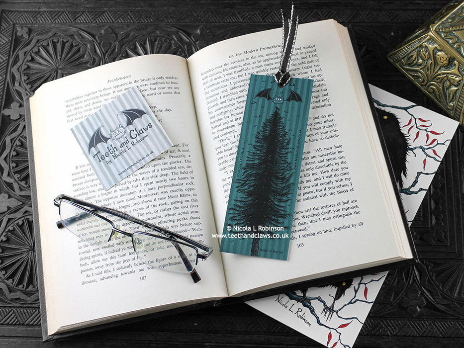 Raven bookmark featuring my original pen and ink illustration, back view of the bookmark resting on an open book with reading glasses and one of my branded cards. Made in the UK. Design and illustration © Nicola L Robinson | Teeth and Claws.