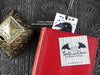 Raven bookmark featuring my original pen and ink illustration, front view with the top showing from inside a closed book with one of my branded cards. Made in the UK. Design and illustration © Nicola L Robinson | Teeth and Claws.