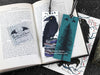 Raven bookmark featuring my original pen and ink illustrations, front and back view of the bookmark resting on an open book with one of my branded cards. Made in the UK. Design and illustration © Nicola L Robinson | Teeth and Claws.