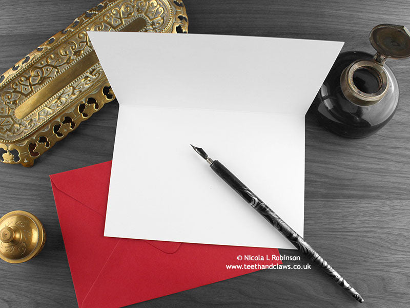Raven and Rose love card, shown open and flat with blank inside, on a red envelope, alongside vintage Victorian pen and writing desk accessories. Design and illustration © Nicola L Robinson | Teeth and Claws