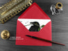 Raven and rose greeting card, shown lying flat, with my original pen and ink illustration of a raven, inside an open red envelope. With vintage Victorian pen and writing desk accessories. Design and illustration © Nicola L Robinson | Teeth and Claws