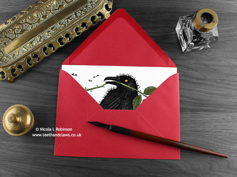 Raven and rose greeting card, shown lying flat, with my original pen and ink illustration of a raven, inside an open red envelope. With vintage Victorian pen and writing desk accessories. Design and illustration © Nicola L Robinson | Teeth and Claws