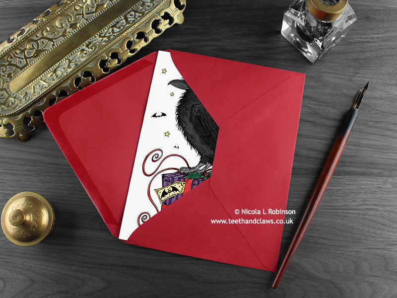 Raven Christmas card, shown lying flat, with my original pen and ink illustration of a festive raven, inside an open red envelope. With vintage Victorian pen and writing desk accessories. Design and illustration © Nicola L Robinson | Teeth and Claws