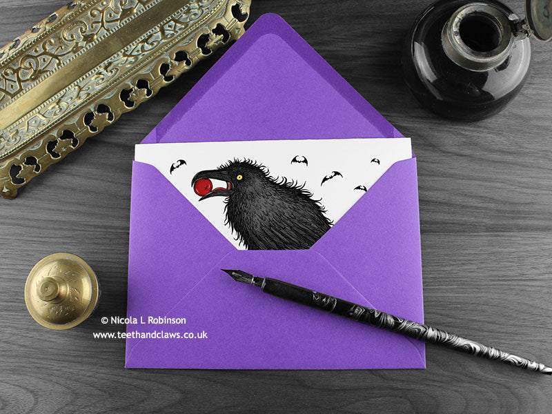 Raven birthday card, shown lying flat, with my original pen and ink illustration of a raven, inside an open purple envelope. With vintage Victorian pen and writing desk accessories. Design and illustration © Nicola L Robinson | Teeth and Claws