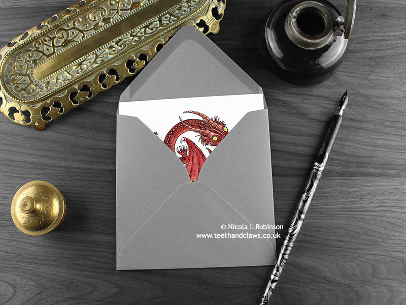 Red Celtic Dragon Greeting Card, shown lying flat, with my original pen and ink illustration of a red dragon, inside an open grey envelope. With vintage Victorian pen and writing desk accessories. Design and illustration © Nicola L Robinson | Teeth and Claws