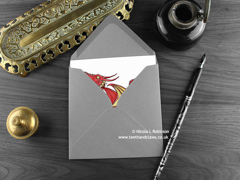 Red Dragon greeting card, shown lying flat, with my original pen and ink illustration of a red dragon, inside an open grey envelope. With vintage Victorian pen and writing desk accessories. Design and illustration © Nicola L Robinson | Teeth and Claws