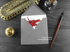 Roaring red dragon greeting card, shown lying flat, with my original pen and ink illustration of a red dragon, inside an open grey envelope. With vintage Victorian pen and writing desk accessories. Design and illustration © Nicola L Robinson | Teeth and Claws