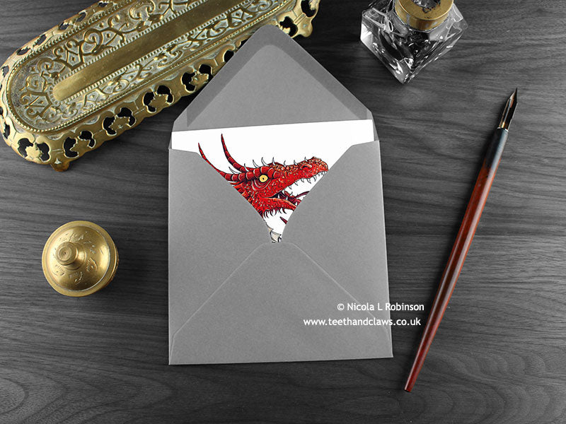 Roaring red dragon greeting card, shown lying flat, with my original pen and ink illustration of a red dragon, inside an open grey envelope. With vintage Victorian pen and writing desk accessories. Design and illustration © Nicola L Robinson | Teeth and Claws