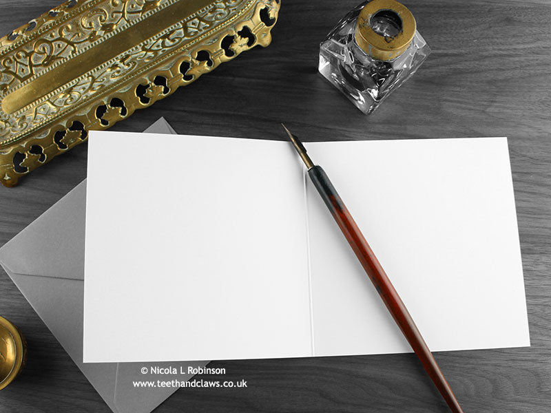 Red fire dragon greeting card, shown open and flat with blank inside, on a grey envelope, alongside vintage Victorian pen and writing desk accessories. Design and illustration © Nicola L Robinson | Teeth and Claws