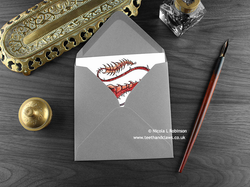 Red fire dragon greeting card, shown lying flat, with my original pen and ink illustration of a red dragon, inside an open grey envelope. With vintage Victorian pen and writing desk accessories. Design and illustration © Nicola L Robinson | Teeth and Claws