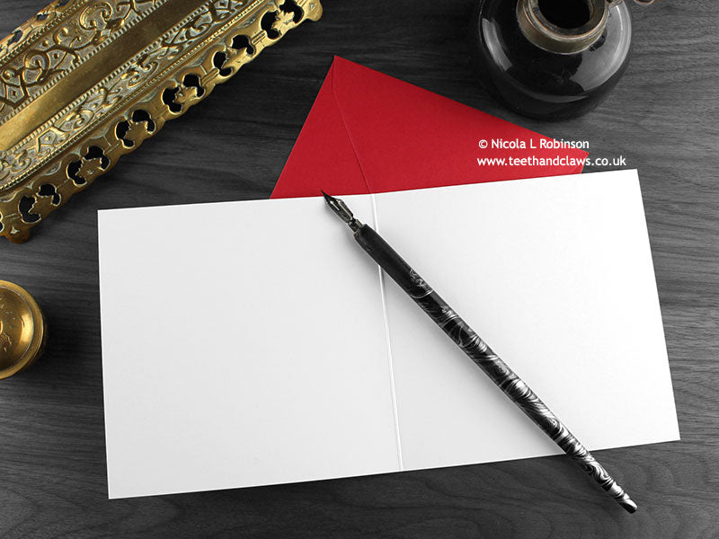 Gothic Reindeer Christmas card, shown open and flat with blank inside, on a red envelope, alongside vintage Victorian pen and writing desk accessories. Design and illustration © Nicola L Robinson | Teeth and Claws