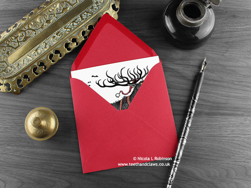 Gothic Reindeer Christmas card, shown lying flat, with my original pen and ink illustration of a Reindeer, inside an open red envelope. With vintage Victorian pen and writing desk accessories. Design and illustration © Nicola L Robinson | Teeth and Claws