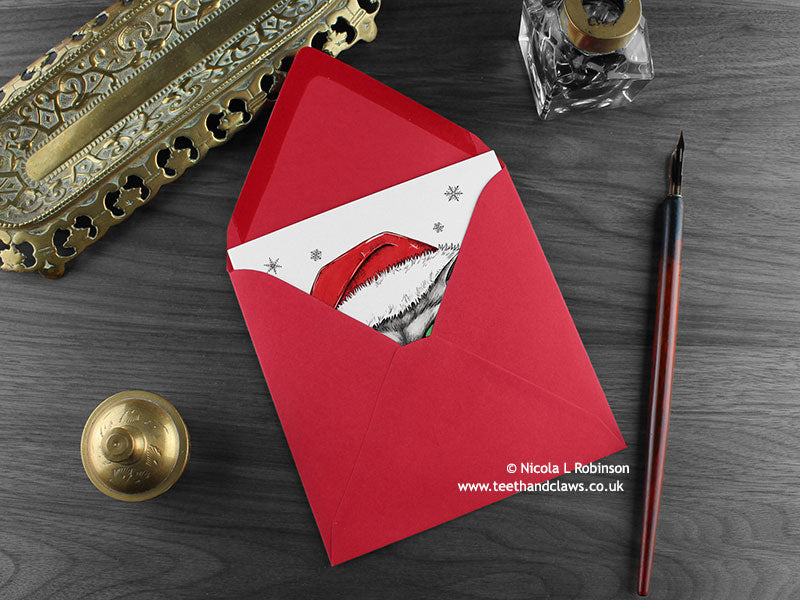 Persian Cat Christmas card, shown lying flat, with my original pen and ink illustration of a festive cat, inside an open red envelope. With vintage Victorian pen and writing desk accessories. Design and illustration © Nicola L Robinson | Teeth and Claws