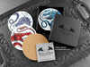 Set of four of my dragon coasters featuring my original pen and ink dragon illustrations, front view alongside a black gift box and card, with one coaster turned over to show the cork backing. Made in the UK. Design and illustration © Nicola L Robinson | Teeth and Claws.