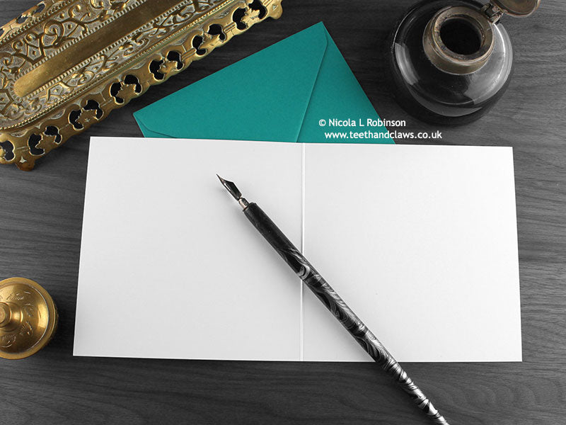 Sorry Cat Greeting card, shown open and flat with blank inside, on a teal envelope, alongside vintage Victorian pen and writing desk accessories. Design and illustration © Nicola L Robinson | Teeth and Claws