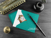 Sorry cat greeting card, shown lying flat, with my original pen and ink illustration of a brown tabby cat, inside an open teal envelope. With vintage Victorian pen and writing desk accessories. Design and illustration © Nicola L Robinson | Teeth and Claws