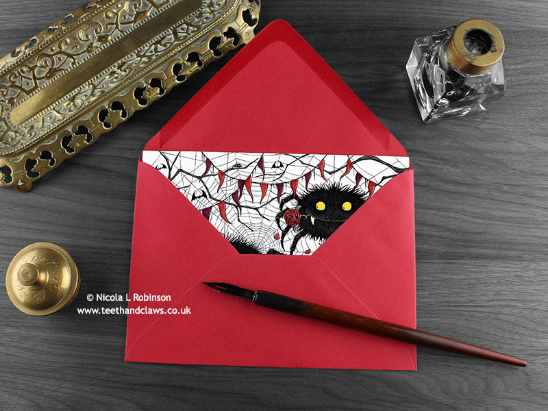 Spider love card, shown lying flat, with my original pen and ink illustration of two spiders, inside an open red envelope. With vintage Victorian pen and writing desk accessories. Design and illustration © Nicola L Robinson | Teeth and Claws