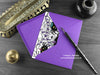 Spider birthday card, shown lying flat, with my original pen and ink illustration of a spider birthday party, inside an open purple envelope. With vintage Victorian pen and writing desk accessories. Design and illustration © Nicola L Robinson | Teeth and Claws