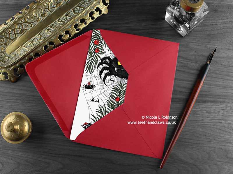 Spider Christmas card, shown lying flat, with my original pen and ink illustration of a festive spider, inside an open red envelope. With vintage Victorian pen and writing desk accessories. Design and illustration © Nicola L Robinson | Teeth and Claws