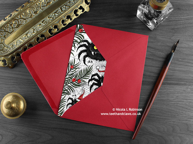 Spiders Christmas card, shown lying flat, with my original pen and ink illustration of festive spiders, inside an open red envelope. With vintage Victorian pen and writing desk accessories. Design and illustration © Nicola L Robinson | Teeth and Claws