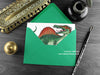 Spinosaurus greeting card, shown lying flat, with my original pen and ink illustration of Spinosaurus, inside an open green envelope. With vintage Victorian pen and writing desk accessories. Design and illustration © Nicola L Robinson | Teeth and Claws
