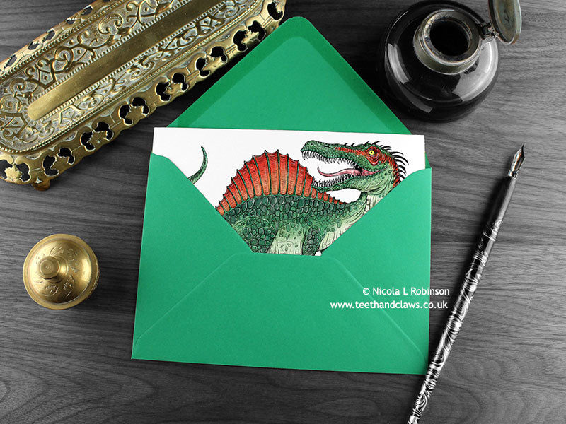 Spinosaurus greeting card, shown lying flat, with my original pen and ink illustration of Spinosaurus, inside an open green envelope. With vintage Victorian pen and writing desk accessories. Design and illustration © Nicola L Robinson | Teeth and Claws