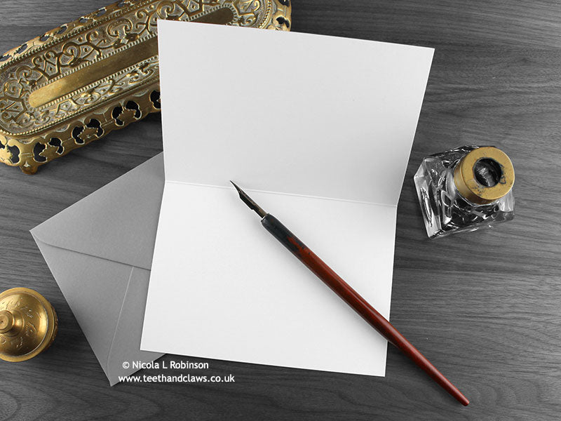 Twin Baby Dragons Greeting Card, shown open and flat with blank inside, on a grey envelope, alongside vintage Victorian pen and writing desk accessories. Design and illustration © Nicola L Robinson | Teeth and Claws