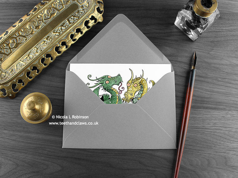 Twin Baby Dragons Greeting Card, shown lying flat, with my original pen and ink illustration of two dragons, inside an open grey envelope. With vintage Victorian pen and writing desk accessories. Design and illustration © Nicola L Robinson | Teeth and Claws