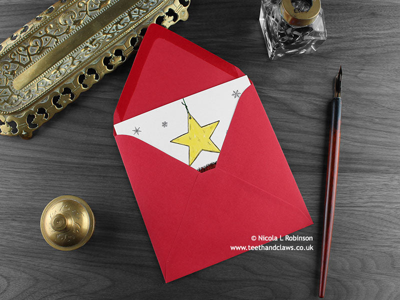 Cat Christmas card, shown lying flat, with my original pen and ink illustration of a festive cat, inside an open red envelope. With vintage Victorian pen and writing desk accessories. Design and illustration © Nicola L Robinson | Teeth and Claws
