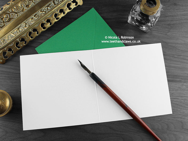 Dinosaur Birthday card, shown open and flat with blank inside, on a green envelope, alongside vintage Victorian pen and writing desk accessories. Design and illustration © Nicola L Robinson | Teeth and Claws