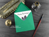 Dinosaur Birthday card, shown lying flat, with my original pen and ink illustration of a Stegosaurus, inside an open green envelope. With vintage Victorian pen and writing desk accessories. Design and illustration © Nicola L Robinson | Teeth and Claws