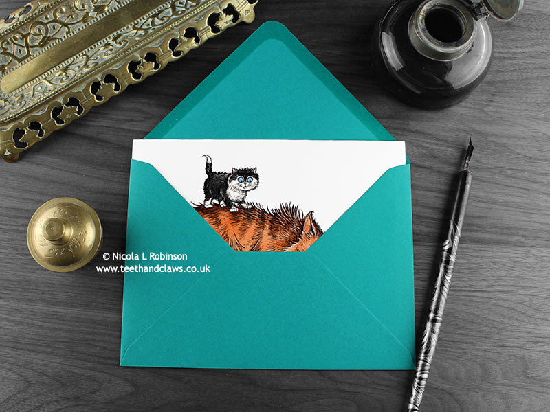 Tom cat and kitten greeting card, shown lying flat, with my original pen and ink illustration of a tom cat and kitten, inside an open teal envelope. With vintage Victorian pen and writing desk accessories. Design and illustration © Nicola L Robinson | Teeth and Claws