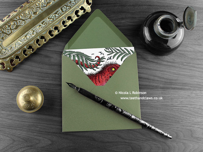 Trex and ferns greeting card, shown lying flat, with my original pen and ink illustration of a dinosaur, inside an open green envelope. With vintage Victorian pen and writing desk accessories. Design and illustration © Nicola L Robinson | Teeth and Claws.