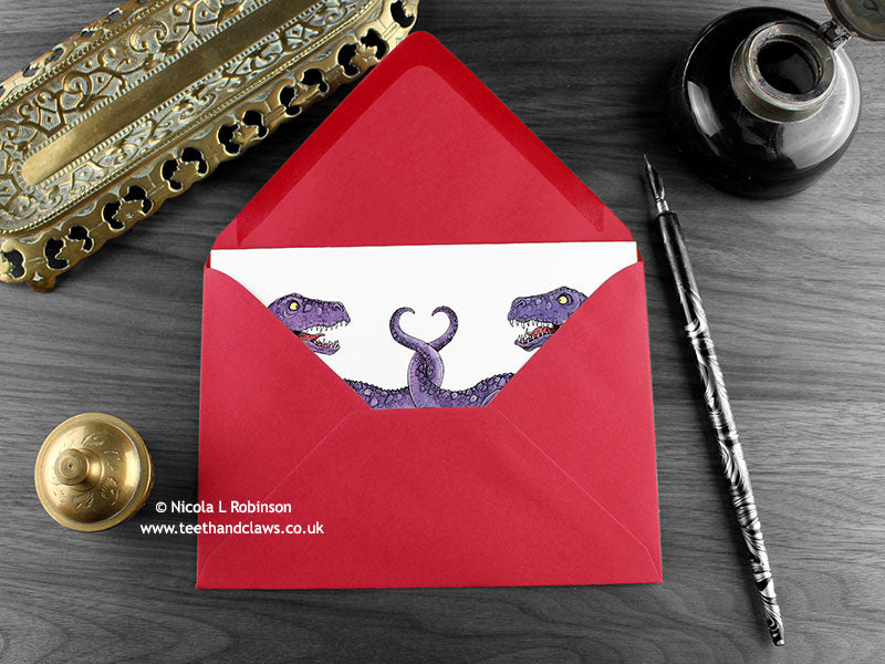 Dinosaur love card, shown lying flat, with my original pen and ink illustration of two dinosaurs, inside an open red envelope. With vintage Victorian pen and writing desk accessories. Design and illustration © Nicola L Robinson | Teeth and Claws