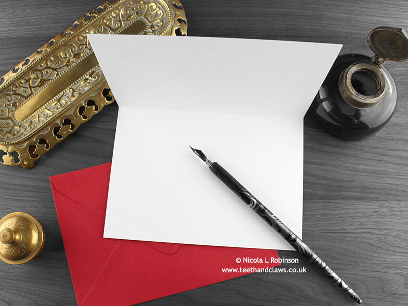 Dinosaur love card, shown open and flat with blank inside, on a red envelope, alongside vintage Victorian pen and writing desk accessories. Design and illustration © Nicola L Robinson | Teeth and Claws