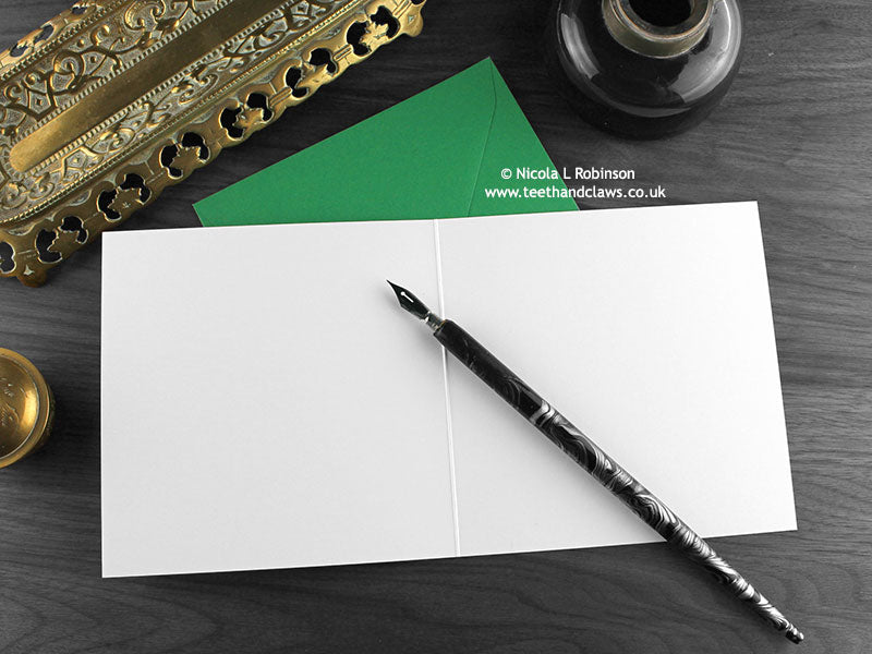 T rex greeting card, shown open and flat with blank inside, on a green envelope, alongside vintage Victorian pen and writing desk accessories. Design and illustration © Nicola L Robinson | Teeth and Claws
