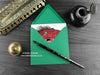 T rex greeting card, shown lying flat, with my original pen and ink illustration of a Tyrannosaurus Rex, inside an open green envelope. With vintage Victorian pen and writing desk accessories. Design and illustration © Nicola L Robinson | Teeth and Claws