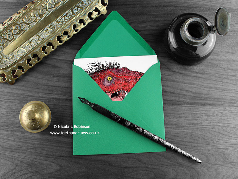 T rex greeting card, shown lying flat, with my original pen and ink illustration of a Tyrannosaurus Rex, inside an open green envelope. With vintage Victorian pen and writing desk accessories. Design and illustration © Nicola L Robinson | Teeth and Claws