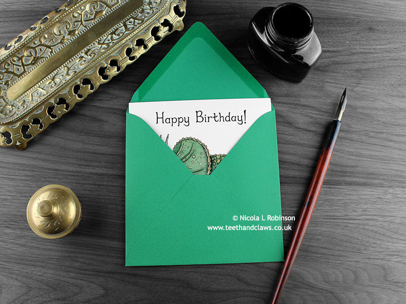 Dinosaur Birthday card, shown lying flat, with my original pen and ink illustration of an Triceratops, inside an open green envelope. With vintage Victorian pen and writing desk accessories. Design and illustration © Nicola L Robinson | Teeth and Claws
