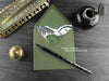Triceratops and ferns greeting card, shown lying flat, with my original pen and ink illustration of a dinosaur, inside an open green envelope. With vintage Victorian pen and writing desk accessories. Design and illustration © Nicola L Robinson | Teeth and Claws.