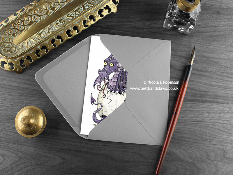 Purple Dragons New Baby Twins Card,, shown lying flat, with my original pen and ink illustration of two dragons, inside an open grey envelope. With vintage Victorian pen and writing desk accessories. Design and illustration © Nicola L Robinson | Teeth and Claws
