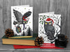 Two Owl Christmas cards, front view from a set of 6, featuring my original pen and ink festive owl illustrations. The cards are standing on books and red envelopes, shown at an angle. Design and illustration © Nicola L Robinson | Teeth and Claws