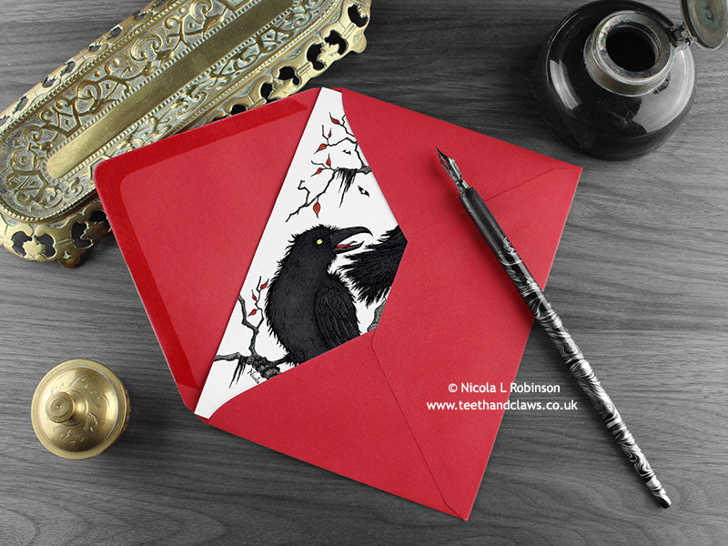 Gothic Ravens love card, shown lying flat, with my original pen and ink illustration of two ravens, inside an open red envelope. With vintage Victorian pen and writing desk accessories. Design and illustration © Nicola L Robinson | Teeth and Claws