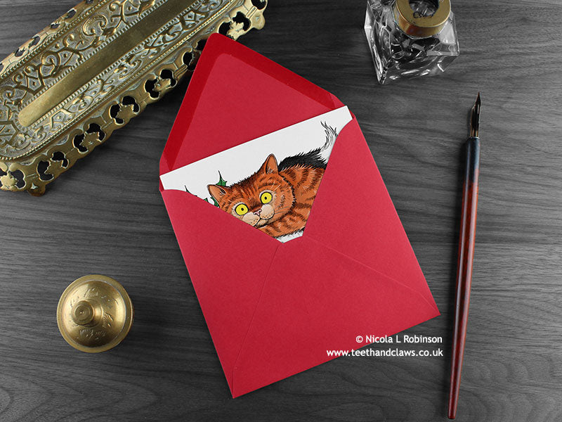 Cats Christmas card, shown lying flat, with my original pen and ink illustration of two festive cats, inside an open red envelope. With vintage Victorian pen and writing desk accessories. Design and illustration © Nicola L Robinson | Teeth and Claws